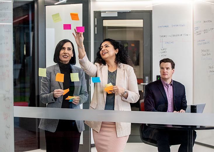 Three online MBA students using colorful sticky notes in a conference room.